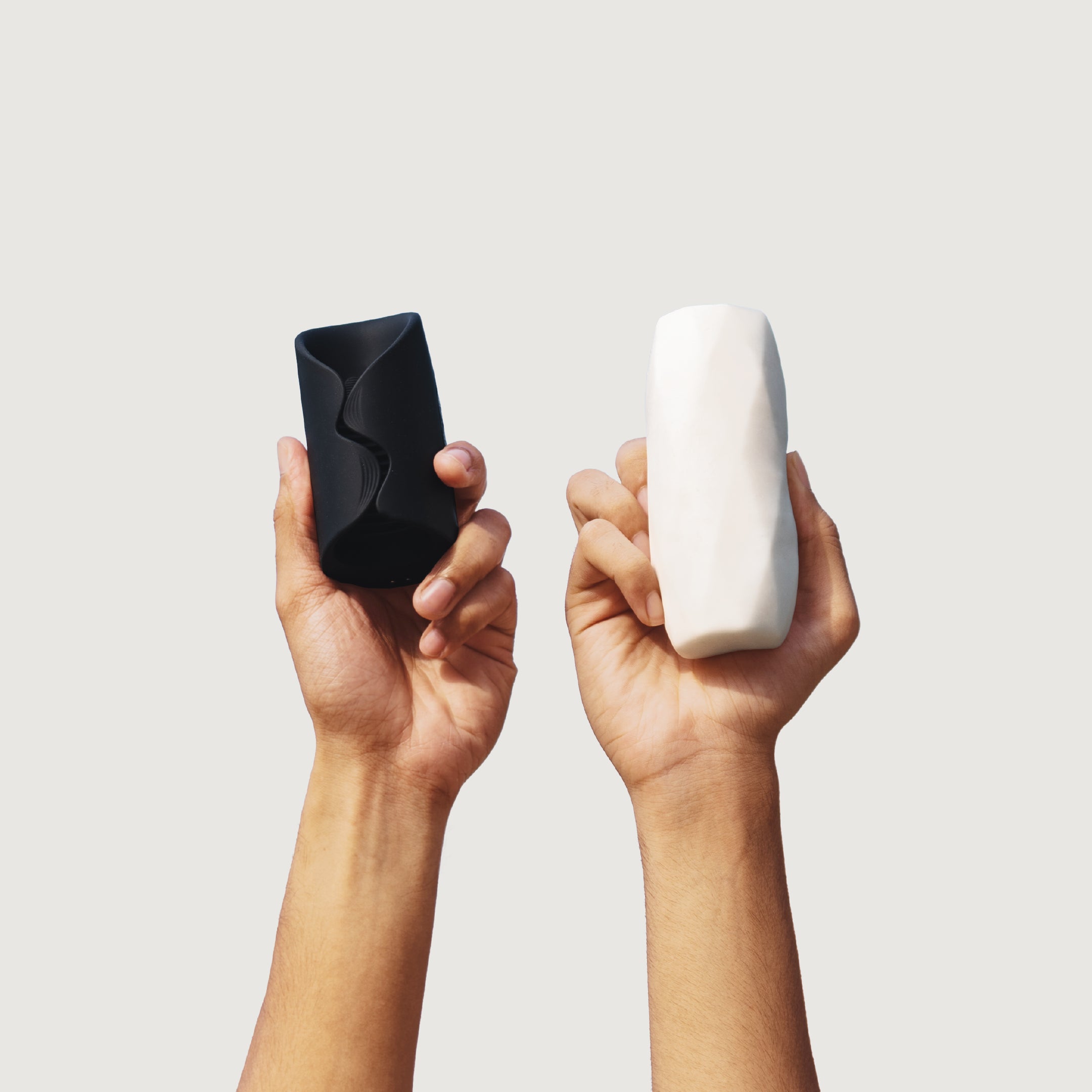 two male hands holding black colored Leo vibrating stroker and white colored Adam manual stroker from Thirdbase held against a white background